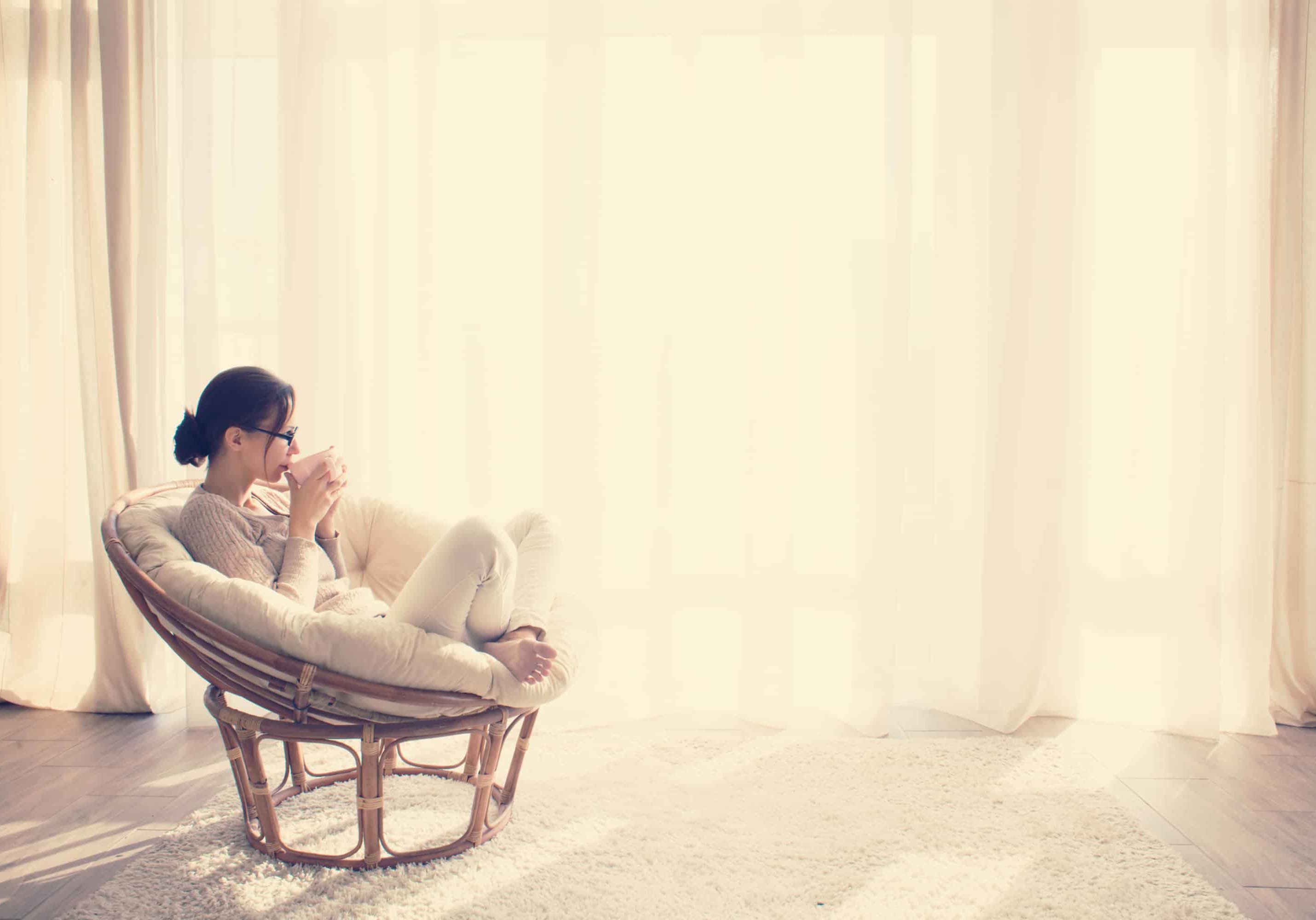 Young woman at home sitting on modern chair in front of window relaxing in her lliving room reading book and drinking coffee or tea